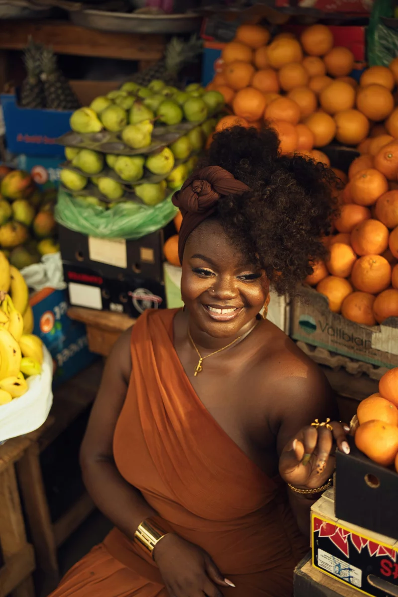 Joyful woman in orange dress surrounded by tropical fruits in an outdoor market.