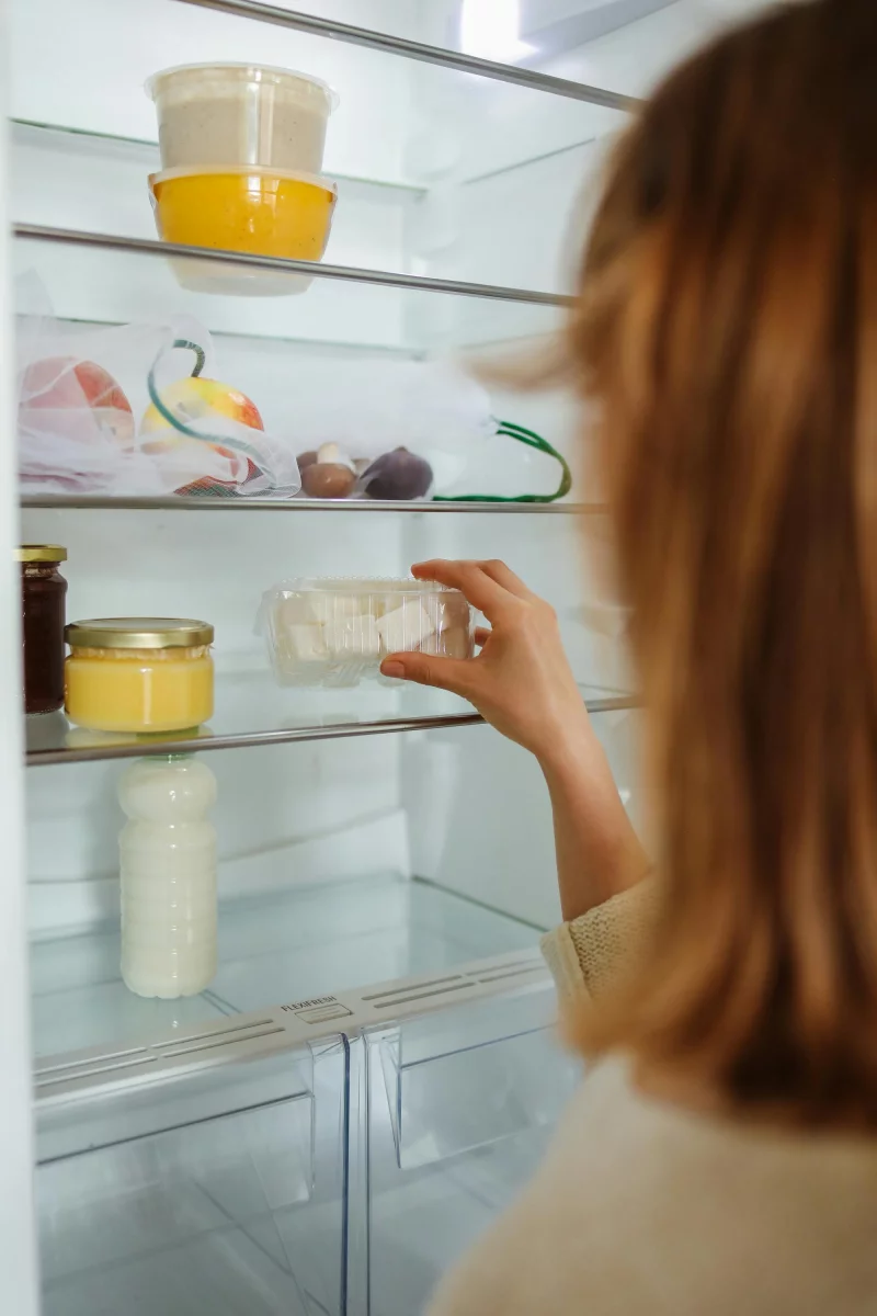 Woman organizing fridge shelves with assorted food containers. Close-up of hand reaching for a jar.