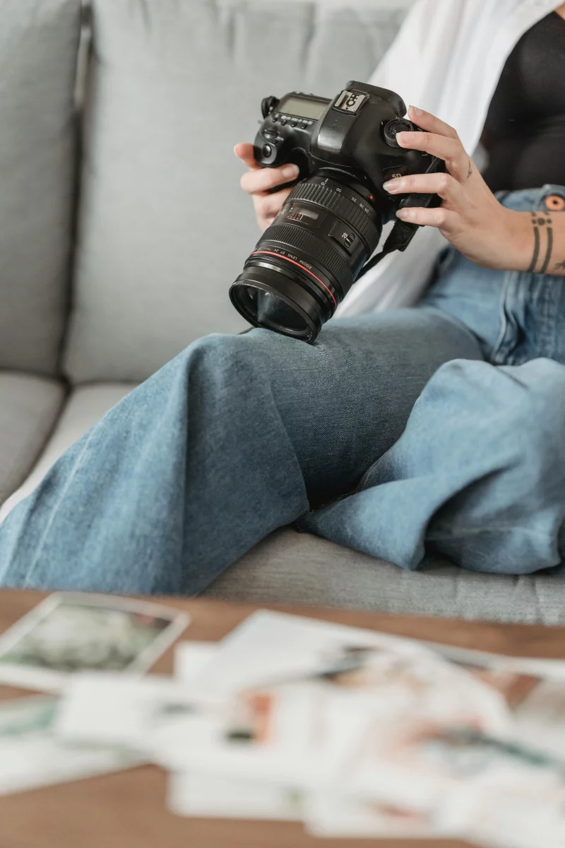A female photographer sits on a sofa indoors reviewing photos on her DSLR camera.