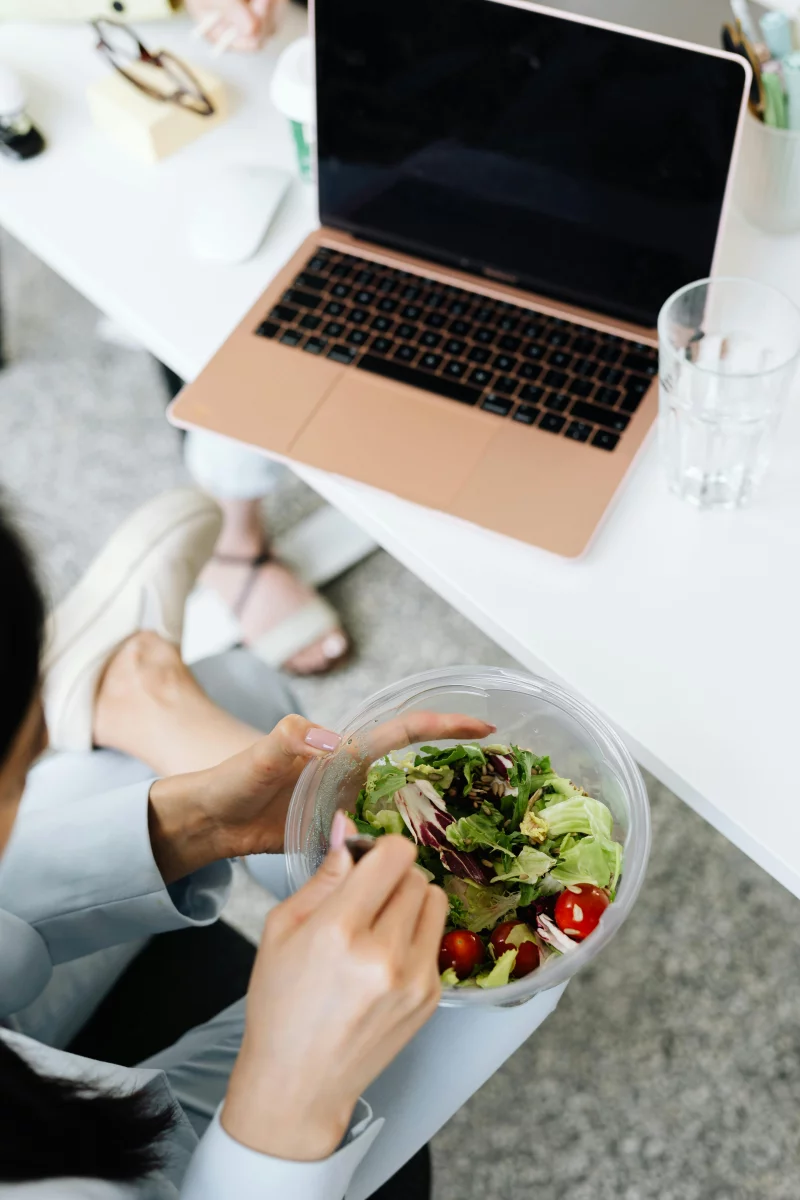 A woman enjoys a healthy salad while working on her laptop in a modern office setting.
