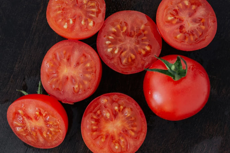 Close-up of fresh ripe tomatoes and slices on a dark wooden surface, showcasing their vibrant red color.