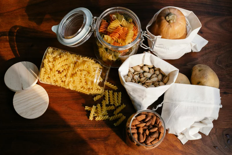 Top view jars of raw pasta placed on wooden table near ECO friendly sacks with pistachios and almonds near pumpkin and potatoes