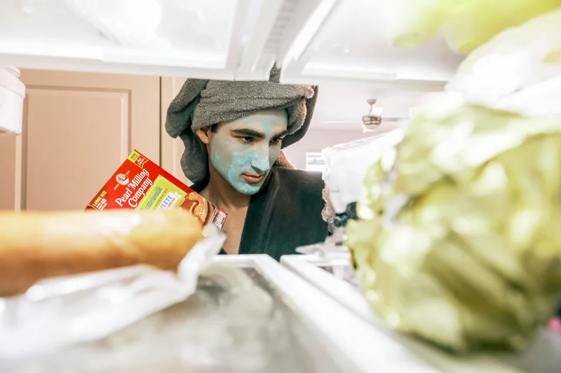 A man with a facial mask and towel looks into a fridge, holding a pancake mix.