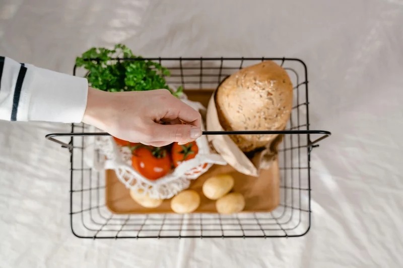 Top view of a hand holding a basket with bread, potatoes, tomatoes, and greens.