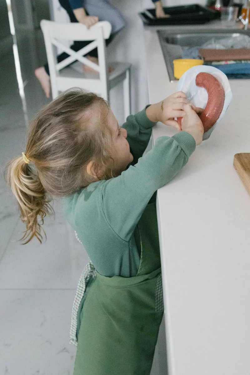 Young child with ponytail reaching for sausage on kitchen counter indoors.