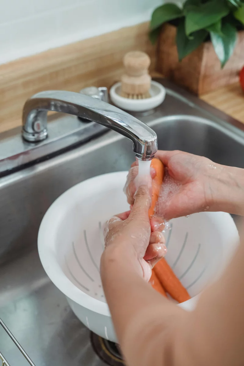 Person washing fresh carrots under a kitchen faucet using a colander, in a home setting.