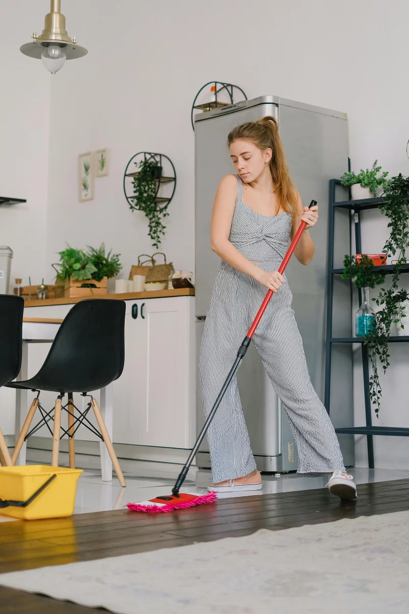 Young carefree female in jumpsuit dancing while cleaning parquet with mop in light house room