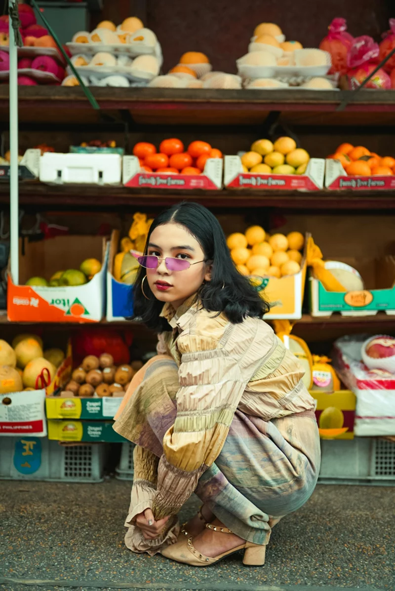 Stylish woman in colorful outfit posing at a lively fruit market stall.