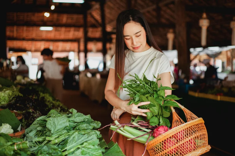 Young ethnic female putting aromatic fresh herb in wicker basket while looking down in local bazaar