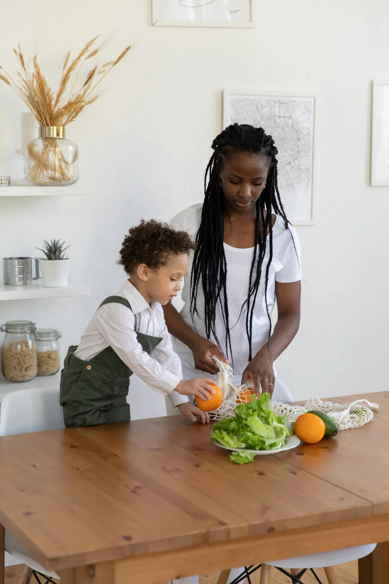 A mother and son standing at a wooden table, arranging fresh fruits and vegetables in a kitchen.