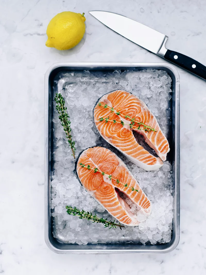 Two fresh salmon steaks seasoned with thyme on ice, accompanied by lemon and knife. Ideal for gourmet recipes.