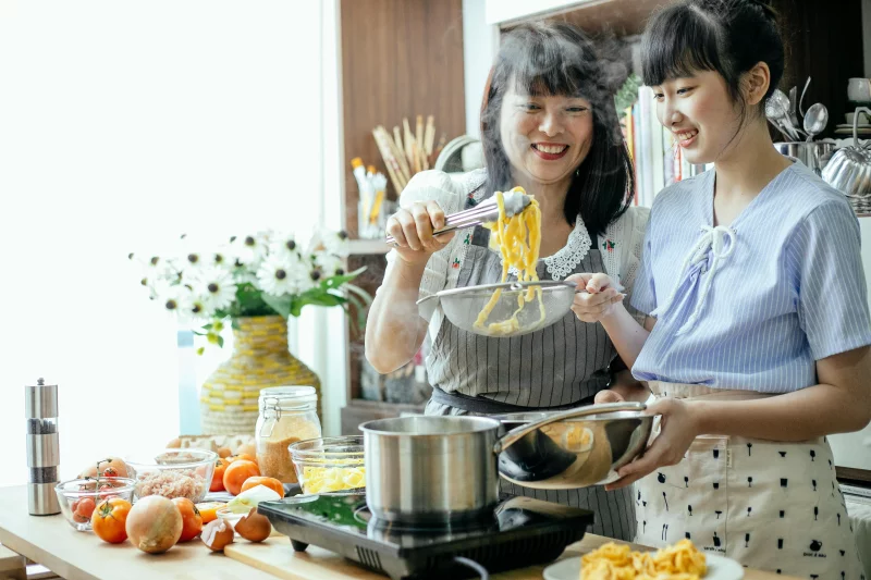 Cheerful Asian teen girl with middle aged mother draining boiled pasta in stainless bowl cooking in team