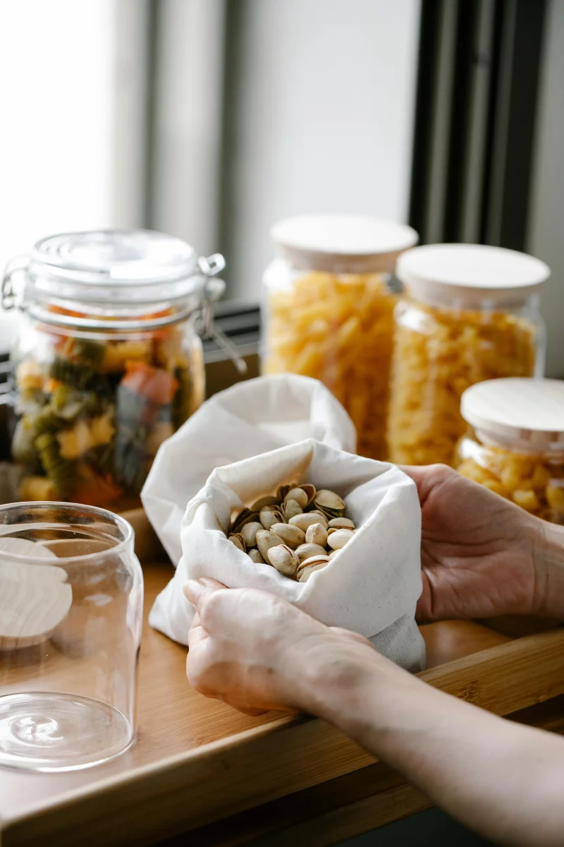 Close-up of hands holding pistachios in a bag next to jars of dried food on a wooden table.