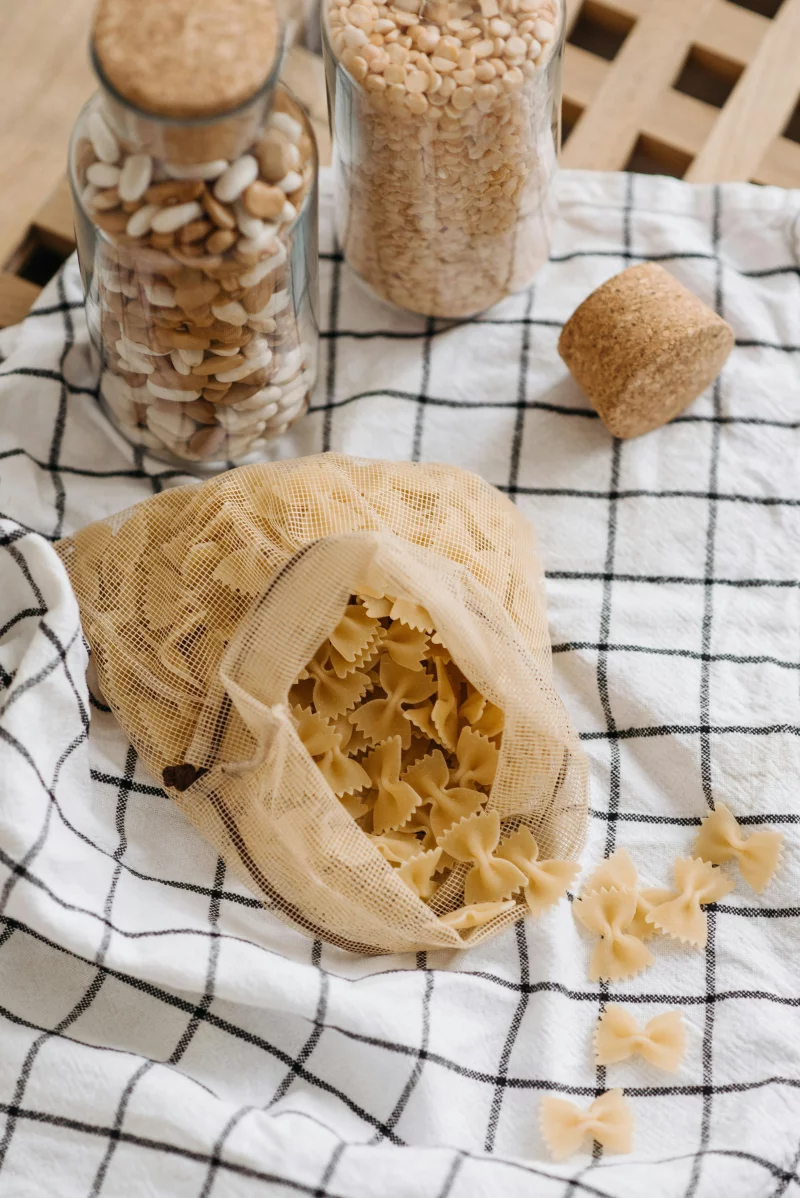 Open eco-friendly bag with uncooked farfalle pasta on a checkered cloth near glass jars.