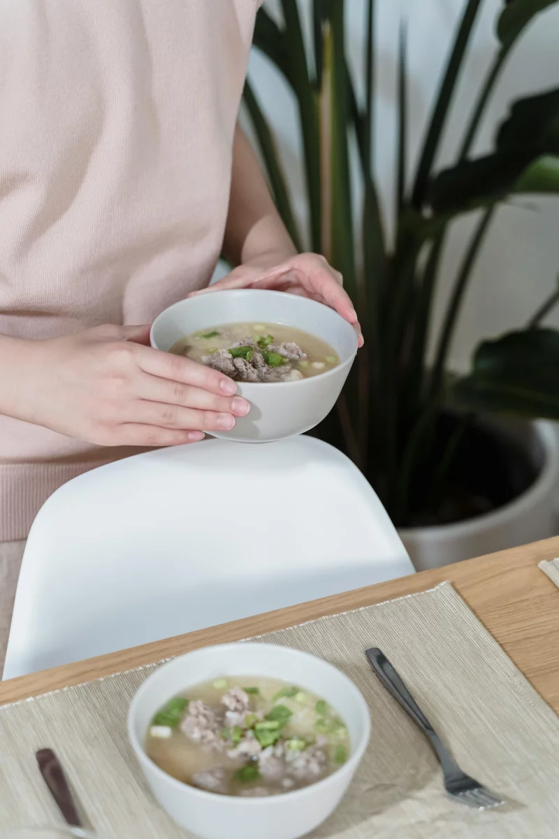 A person holding a bowl of hot soup at a dining table, with a focus on fresh ingredients.