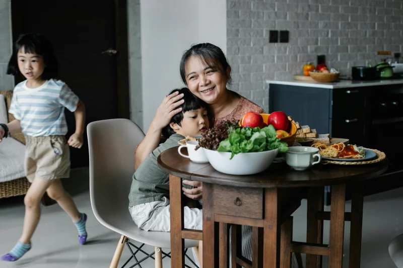 Happy kind ethnic senior female hugging cute grandson having breakfast in kitchen while active little granddaughter running behind table