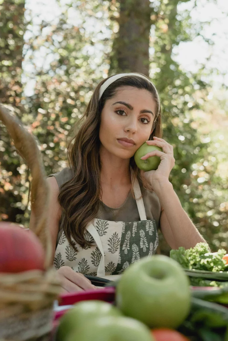 A woman holding a green apple while sitting at a table surrounded by fresh fruit and vegetables in a garden.