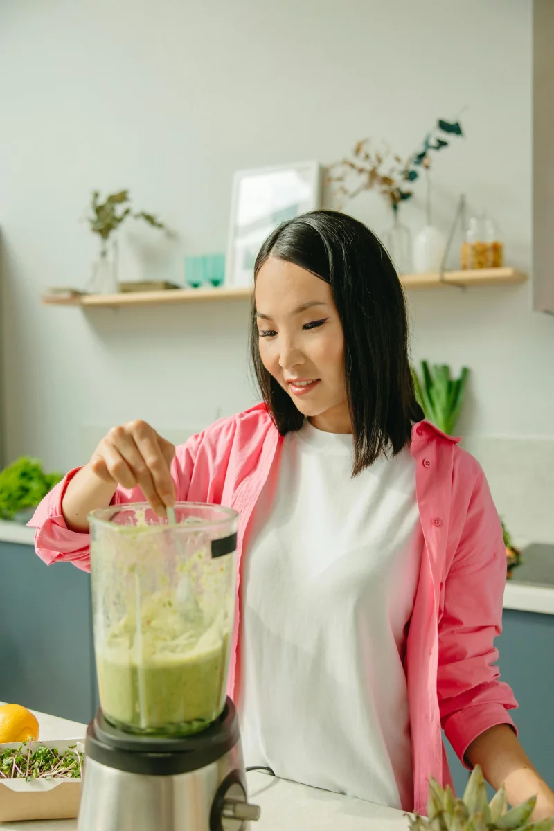 Asian woman making a healthy green smoothie in a modern kitchen setting.