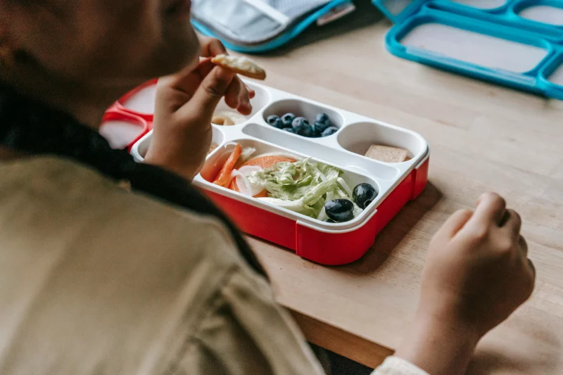 Crop faceless teenage sitting at wooden table with lunch box full of healthy food