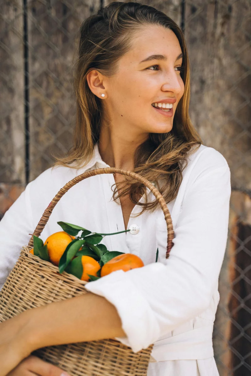 A cheerful woman in white holds a woven basket filled with fresh tangerines, radiating joy.