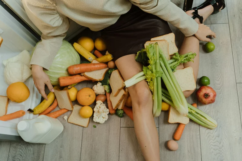 A woman amidst spilled groceries, highlighting food waste and consumerism with vegetables, fruits, and bread.