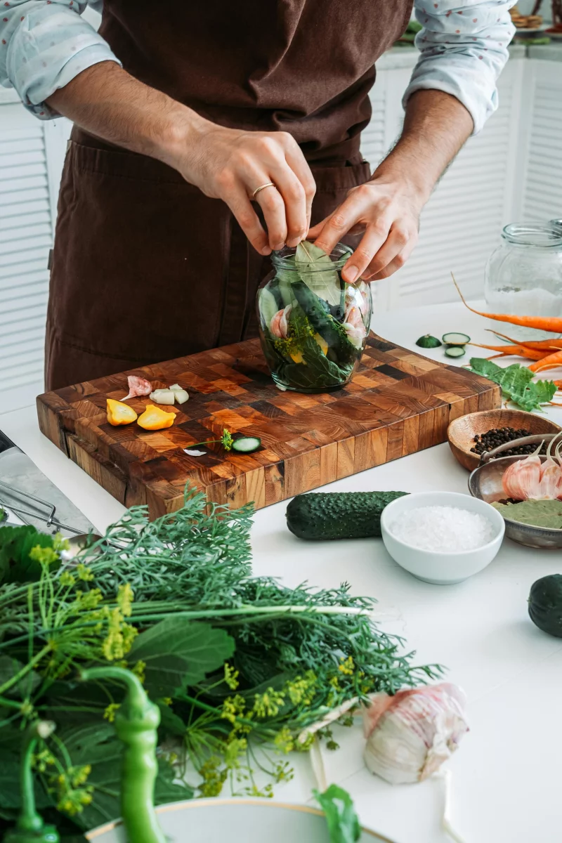 A man prepares fresh herbs and vegetables for pickling indoors on a wooden board.