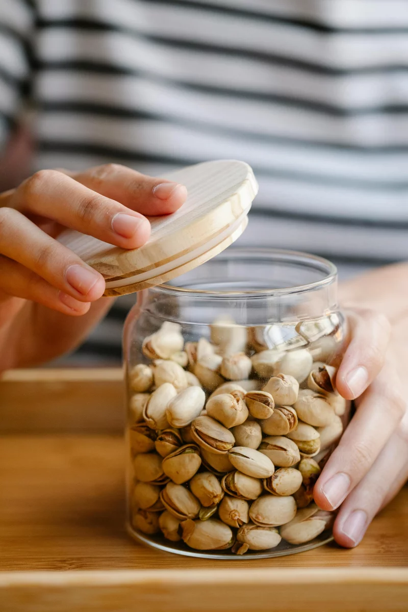 Close-up of hands holding a glass jar of pistachios with a wooden lid indoors.