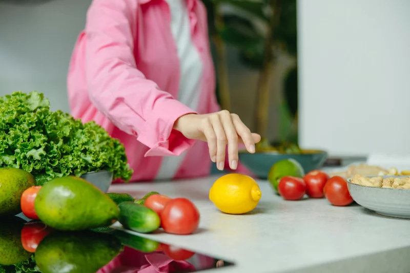 A person in a pink shirt reaching for a lemon surrounded by fresh vegetables.