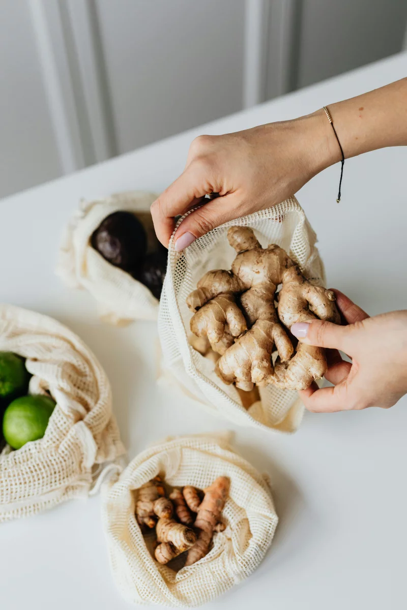 Hands placing ginger into an eco-friendly reusable net bag on a kitchen counter.