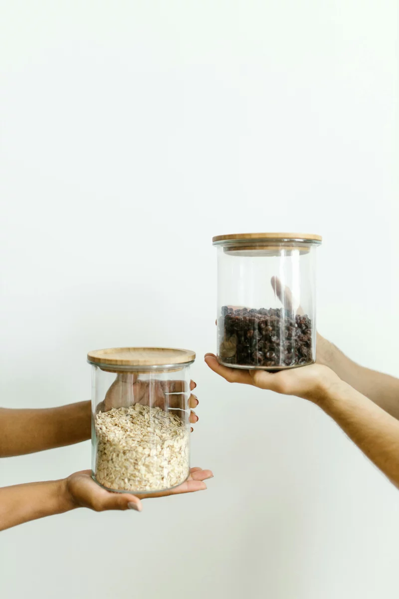 Two hands hold glass jars filled with grains and beans against a white wall.