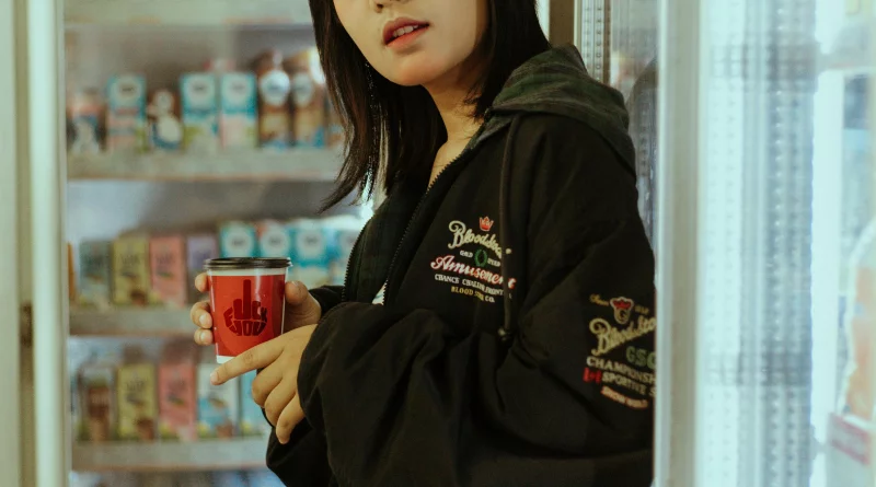 Asian woman holding a vibrant coffee cup indoors, wearing a black jacket.