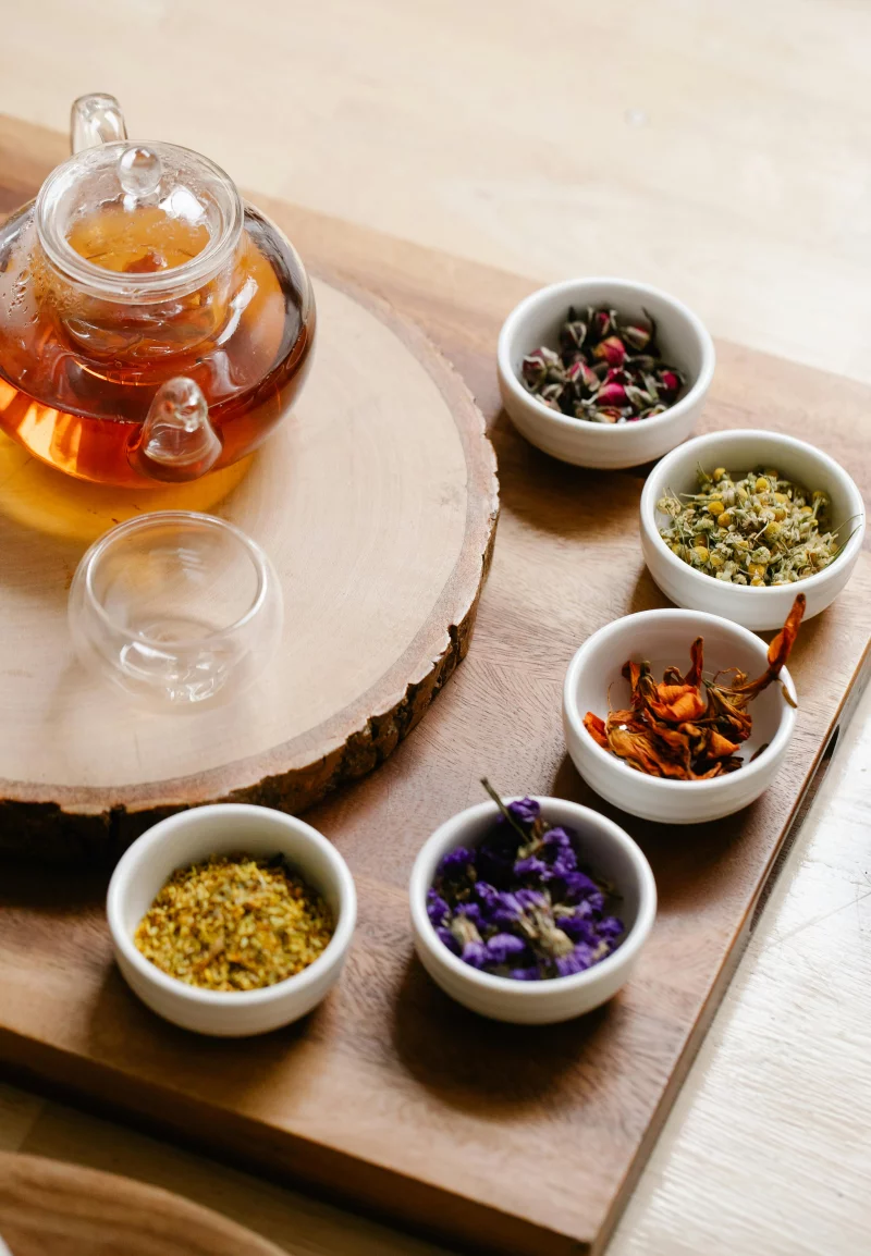Glass teapot with herbal tea and assorted dry herbs in bowls on a wooden surface for aromatherapy.