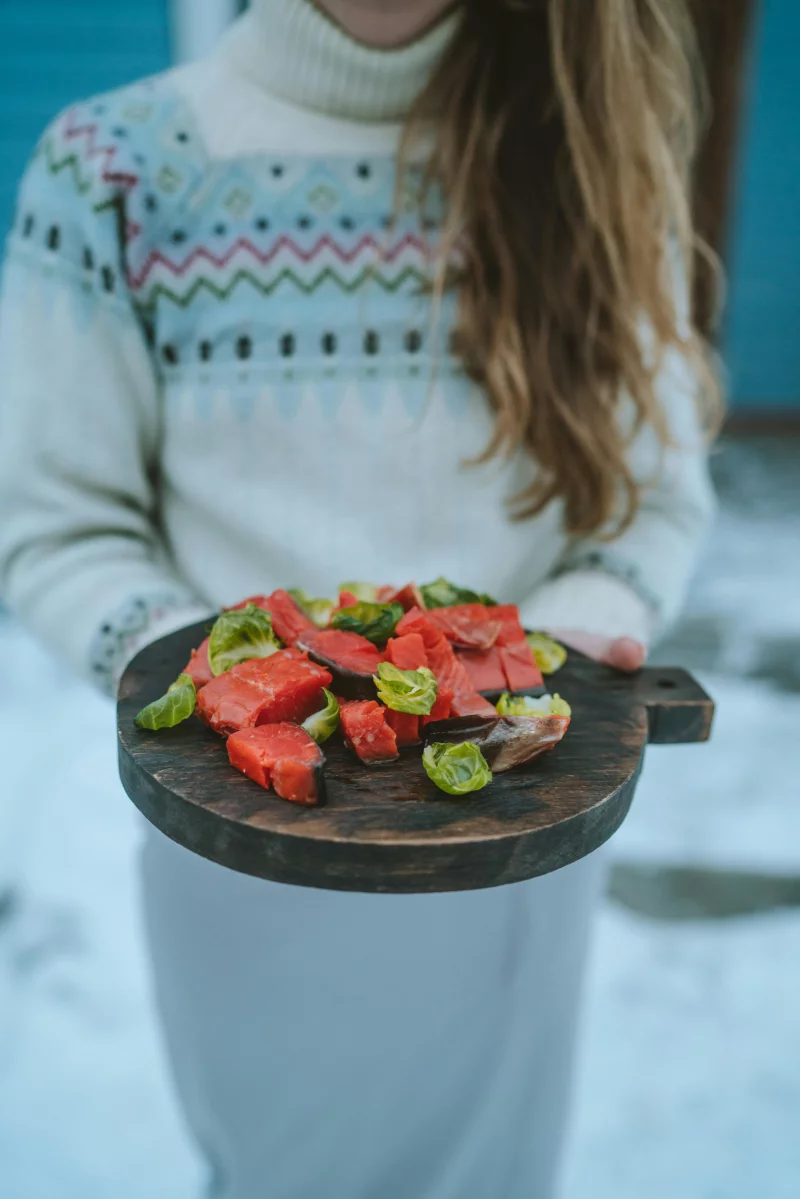 A woman holds a wooden platter with salmon and Brussels sprouts in a snowy outdoor setting.