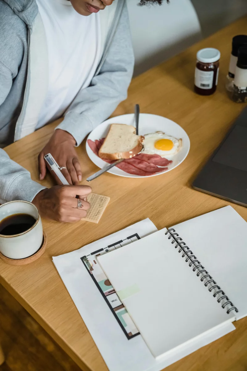 A woman writes notes beside a breakfast plate with toast, eggs, and meats on a wooden table.