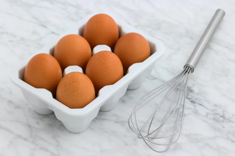 Six brown eggs in a carton and a metal whisk on a marble countertop. Perfect for cooking concepts.