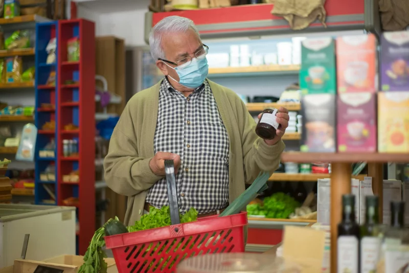 Elderly man with mask shopping for groceries indoors, carrying a basket.
