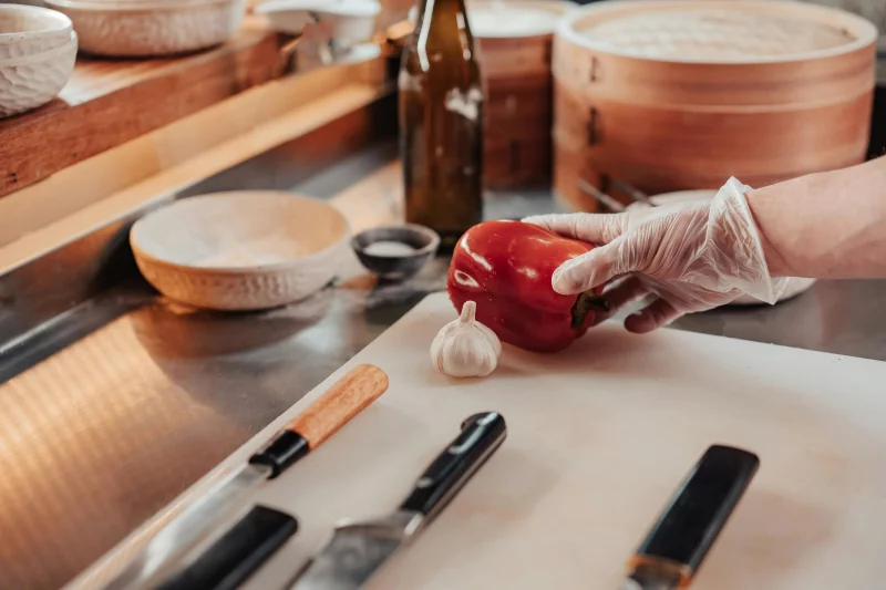 A chef wearing gloves prepares a bell pepper on a cutting board amidst modern kitchen tools.