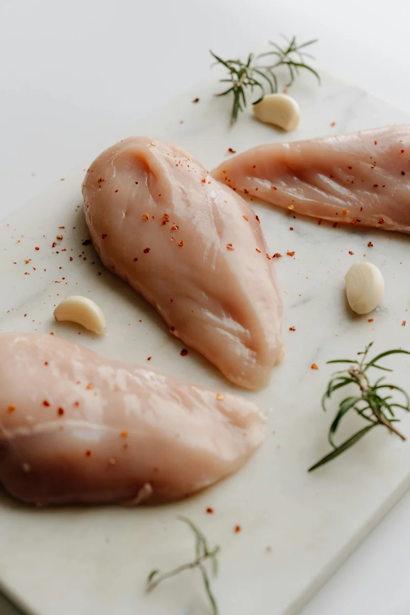 Three raw chicken breasts seasoned with herbs and whole garlic on a marble surface.