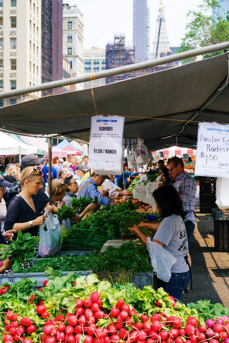 Lively city market scene with people buying fresh vegetables at outdoor stalls.
