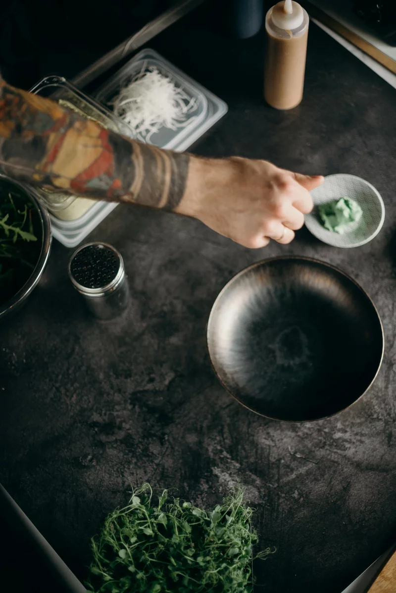 Tattooed chef prepares fresh greens and ingredients in a moody kitchen setup.