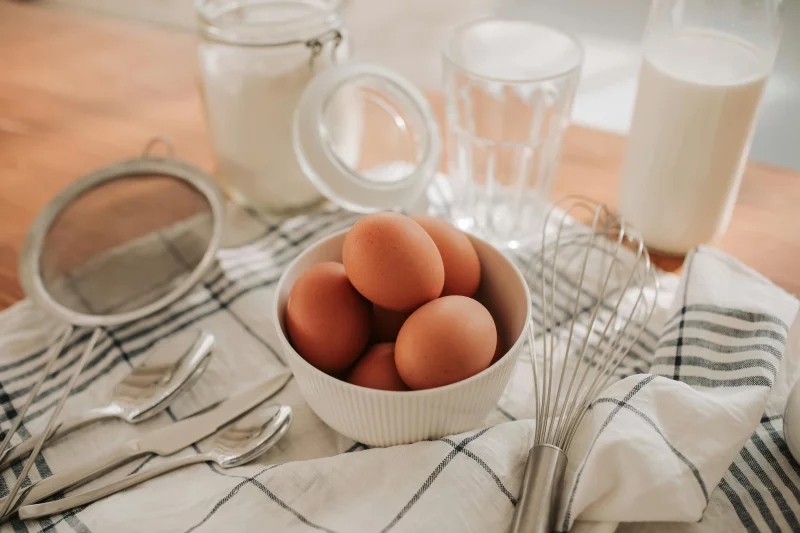 Close-up of fresh eggs in a bowl with kitchen utensils on a striped cloth for cooking preparation.