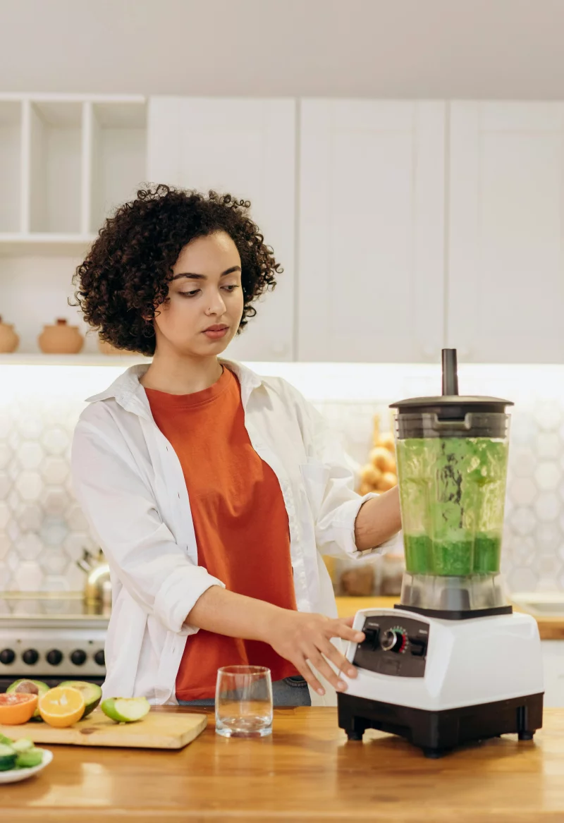 Curly haired woman preparing a green smoothie in a modern kitchen setting.