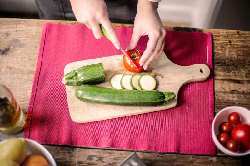 Hands slicing tomato and zucchini on a wooden board for a vibrant kitchen scene.