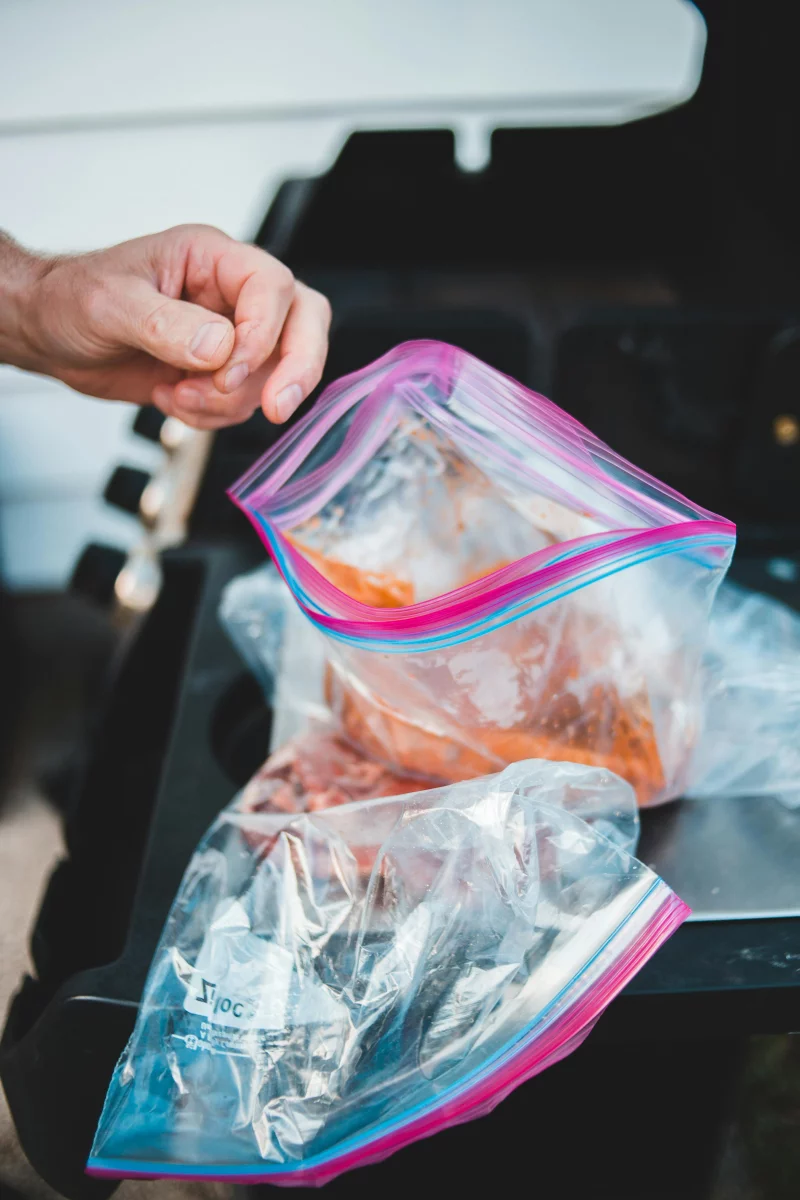 Close-up of hand preparing marinated meat in a Ziploc bag on a grill.