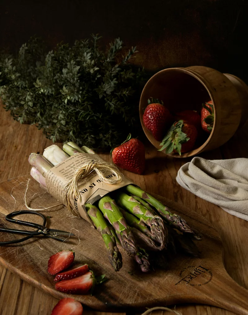 A dark, rustic still life with asparagus and strawberries on a wooden surface.