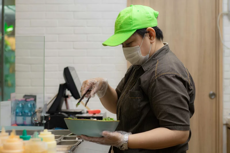 A chef in a commercial kitchen prepares a salad using tongs and a bowl.