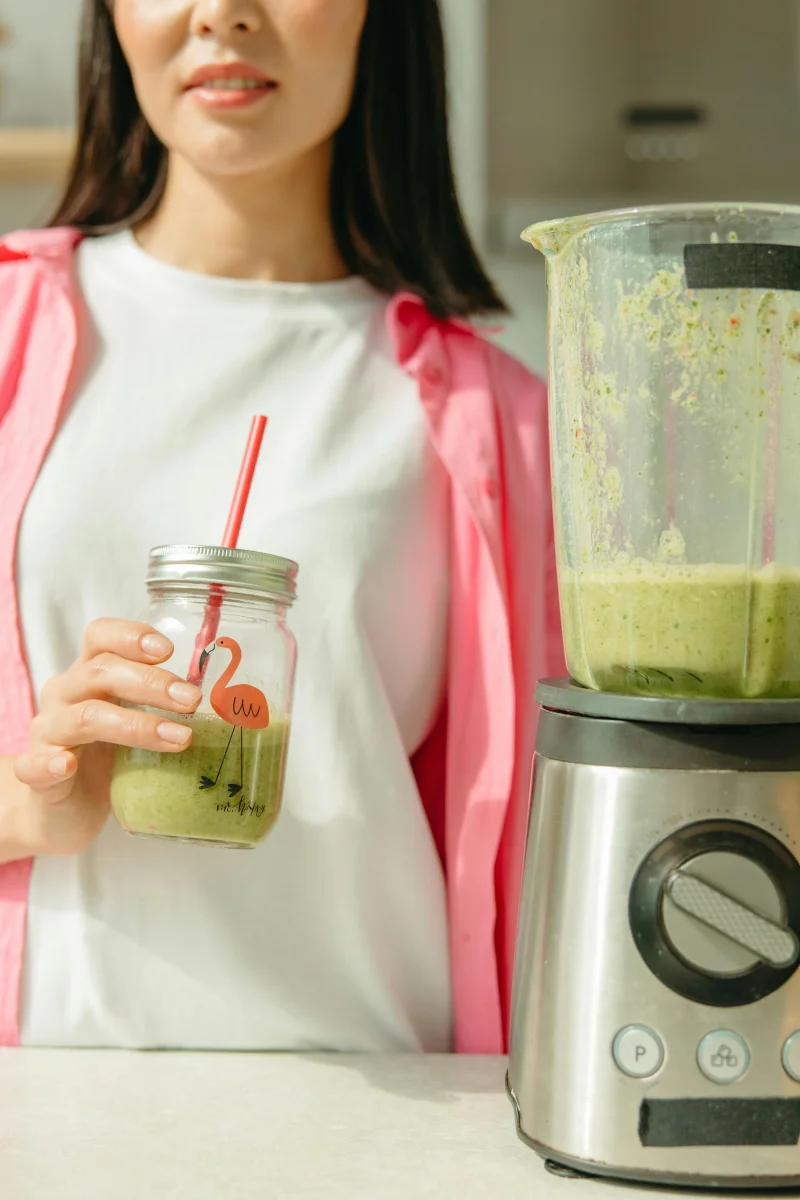 A woman in a pink shirt holding a healthy green smoothie in a kitchen setting.