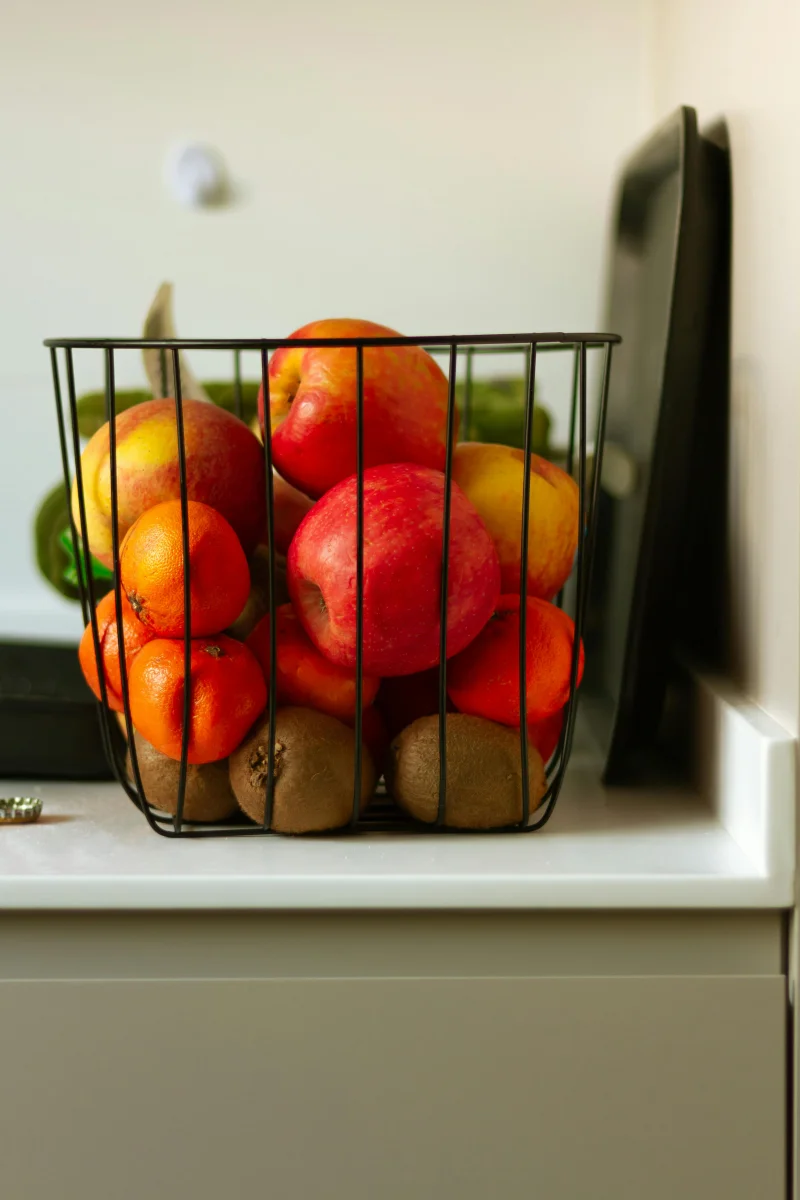 Assorted fresh apples, mandarins, and kiwis in a wire basket on kitchen counter.