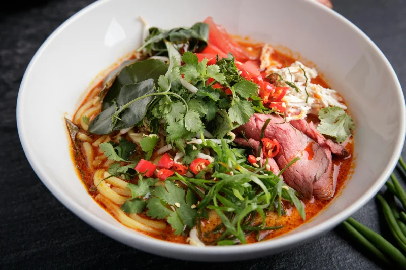 A vibrant and spicy Asian noodle soup featuring fresh herbs, beef, and chili peppers in a white bowl.