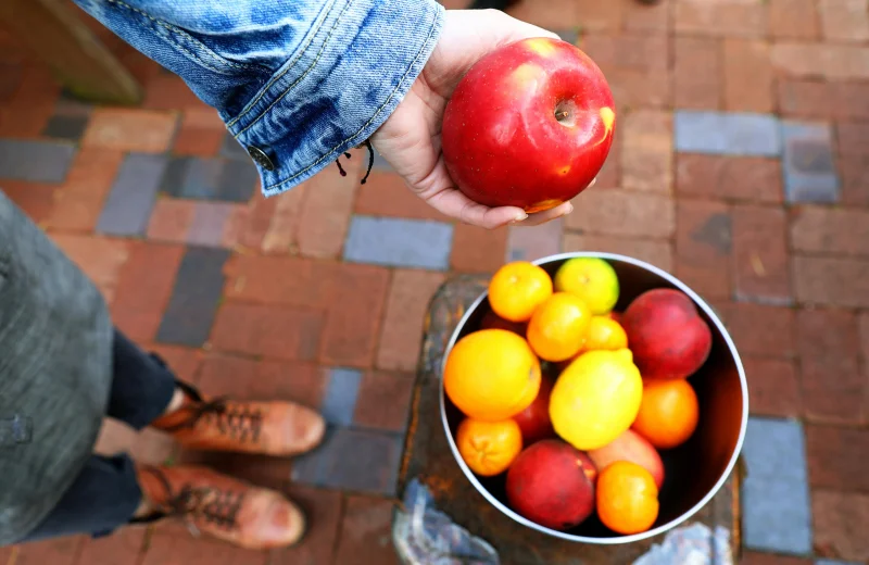 Top view of a person holding a red apple above a bowl of fresh fruits on a brick surface.